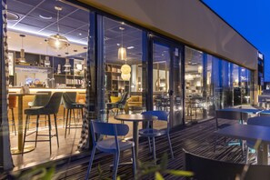 Un espace de détente intérieur/extérieur avec des tables et des chaises, un coin bar avec des étagères et des bouteilles, et une vue à travers de grandes portes vitrées.