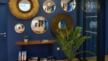 A room with a blue wall, a wooden console table with magazines, a large potted plant, and multiple mirrors.