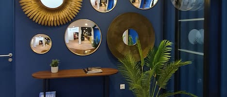 Una habitación con pared azul, una consola de madera con revistas, una planta grande en maceta y varios espejos.
