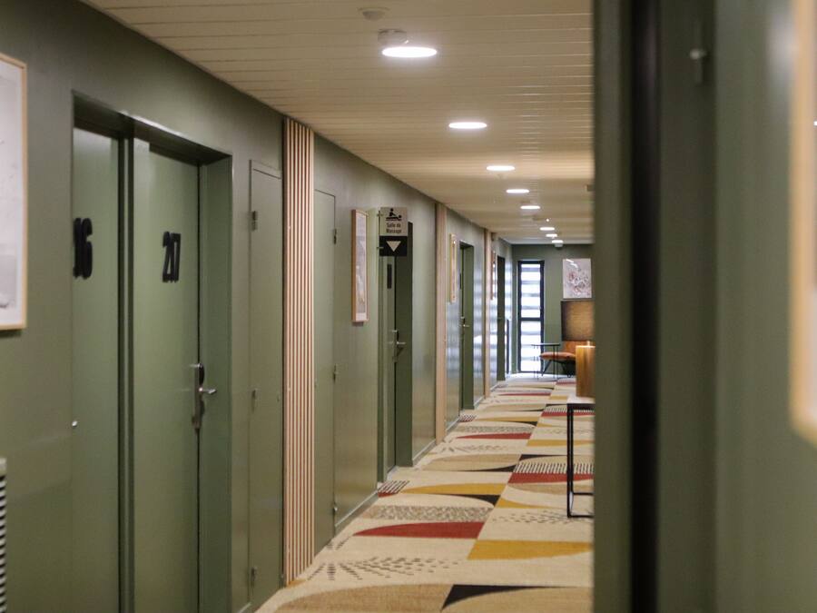 Un pasillo con puertas numeradas del 16 al 21, un cartel de indicaciones y una alfombra estampada.