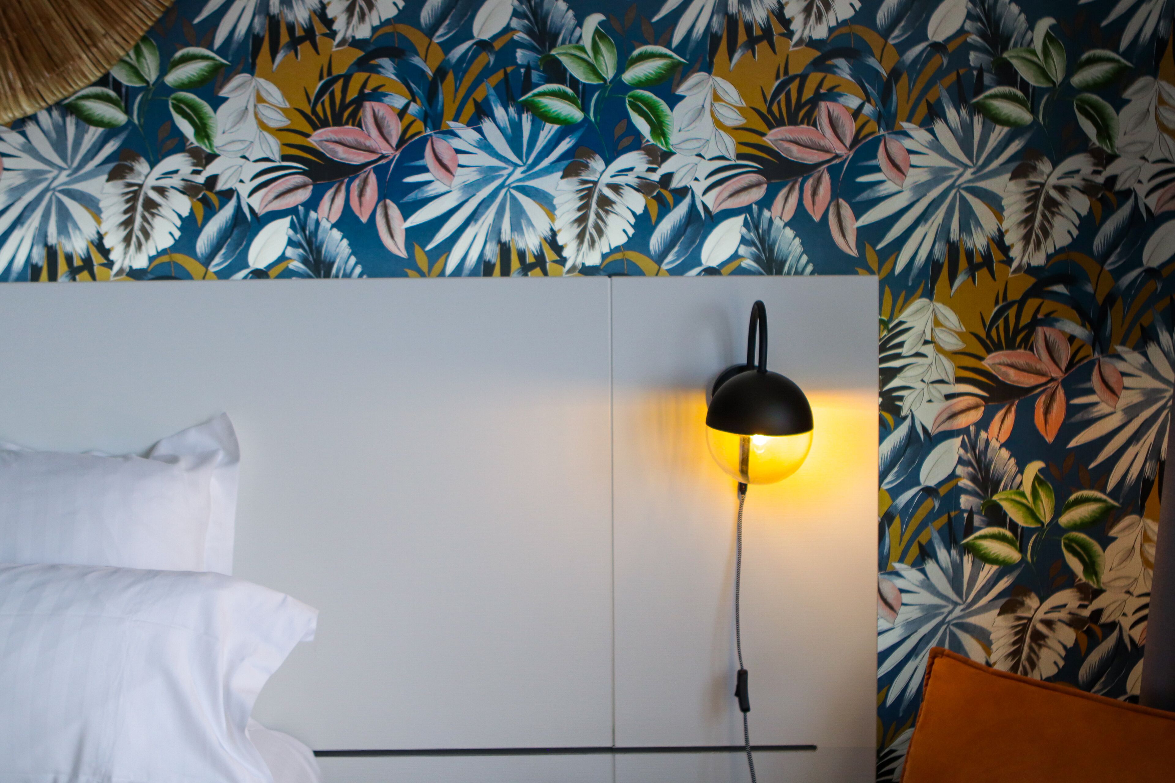 Un dormitorio con cama, mesita de noche, lámpara de pared y papel tapiz con estampado floral.