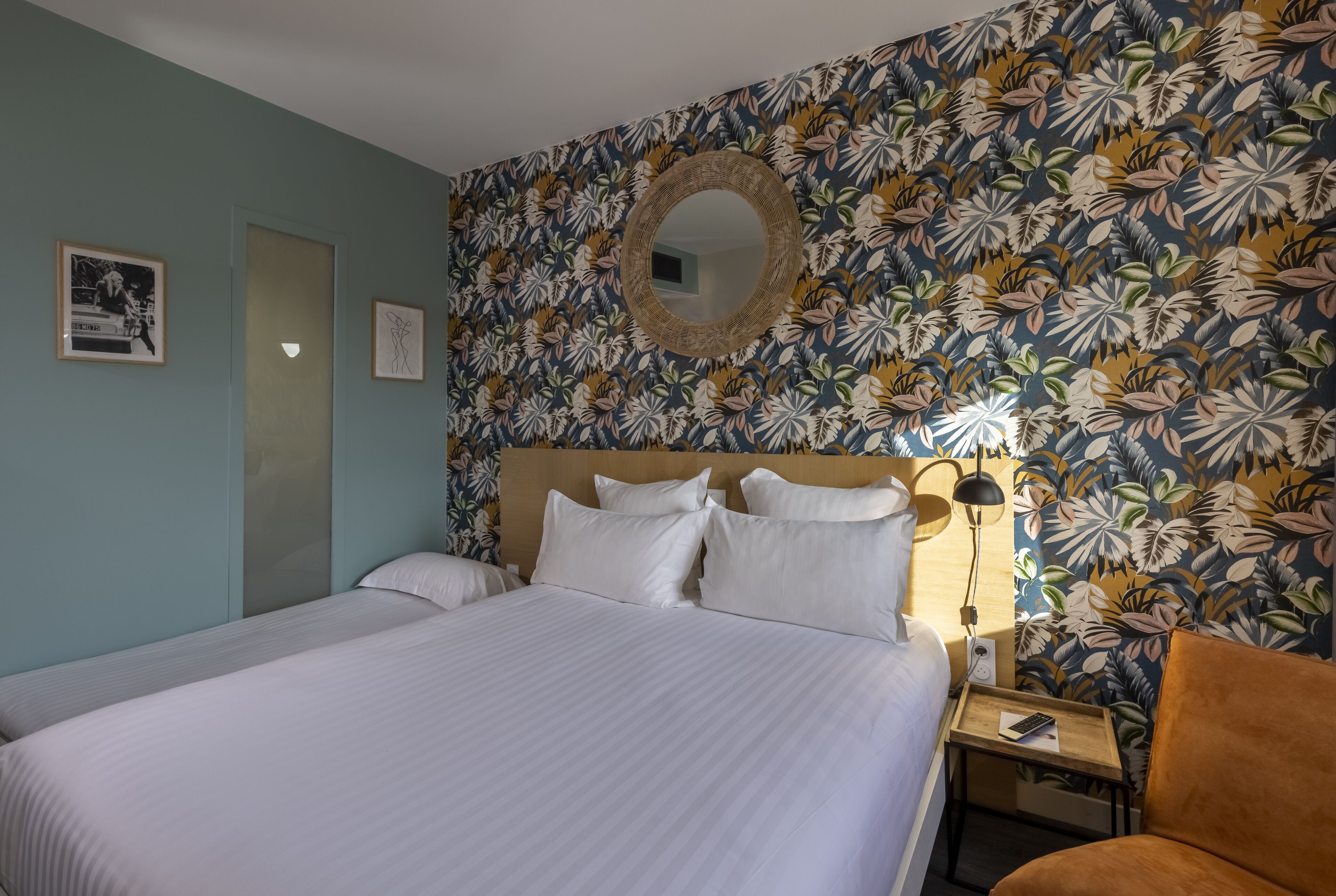 A neatly made bed with white linens, a wooden headboard, and a wall with a tropical leaf pattern. There is a round mirror above the bed and a small bedside table with a lamp and a remote control.
