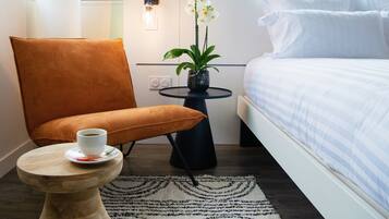 Una camera d'albergo con un letto, una sedia, un tavolino con una tazza, un'orchidea in vaso e una lampada a parete.