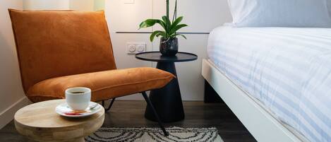 Habitación de hotel con cama, una silla, una mesita con una taza, una orquídea en maceta y una lámpara de pared.