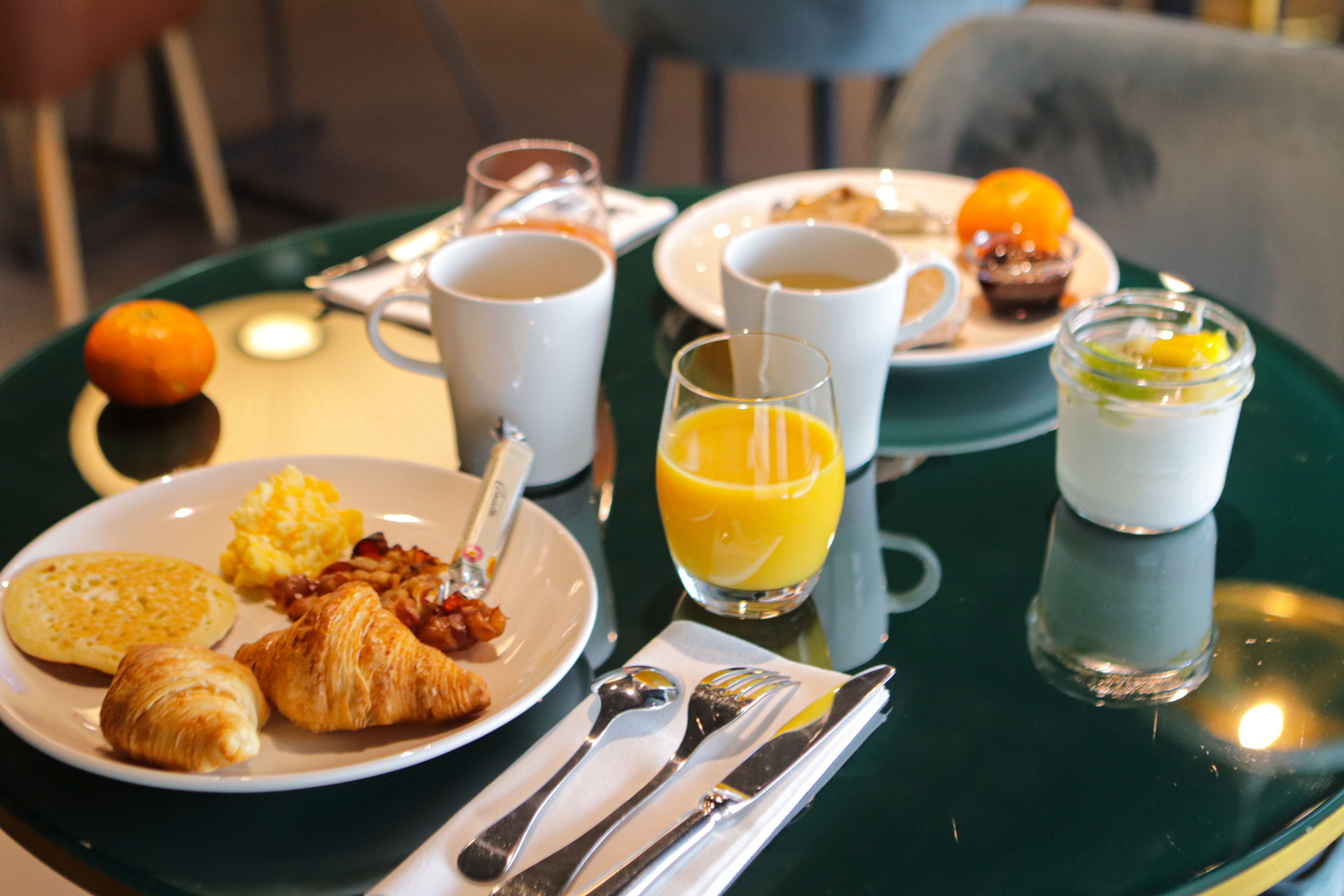Une table ronde dressée pour le petit-déjeuner, avec des assiettes remplies de nourriture, des tasses et un verre de jus d’orange.
