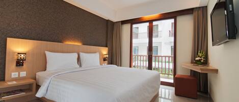 A modern hotel room with a large bed, a nightstand with a lamp, a TV mounted on the wall, and a balcony with a view of the building.