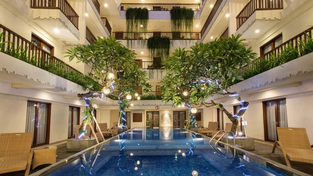 A multi-level indoor swimming pool with a central water feature, surrounded by lounge areas and balconies with plants. - Sense Sunset Hotel Seminyak (Seminyak)