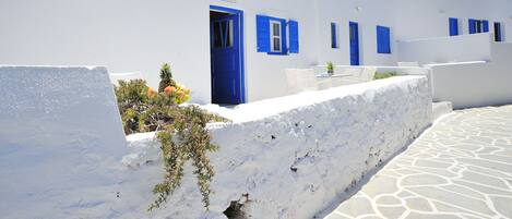 A white building with blue doors and windows, a stone pathway, and a small garden with plants and a tree.