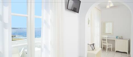 A bright room with a TV, a desk, a chair, a table with a glass, and a view of the sea.