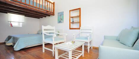 Une pièce avec un lit en mezzanine, deux chaises blanches avec des coussins bleus, une petite table, un canapé bleu clair et une bibliothèque en bois.