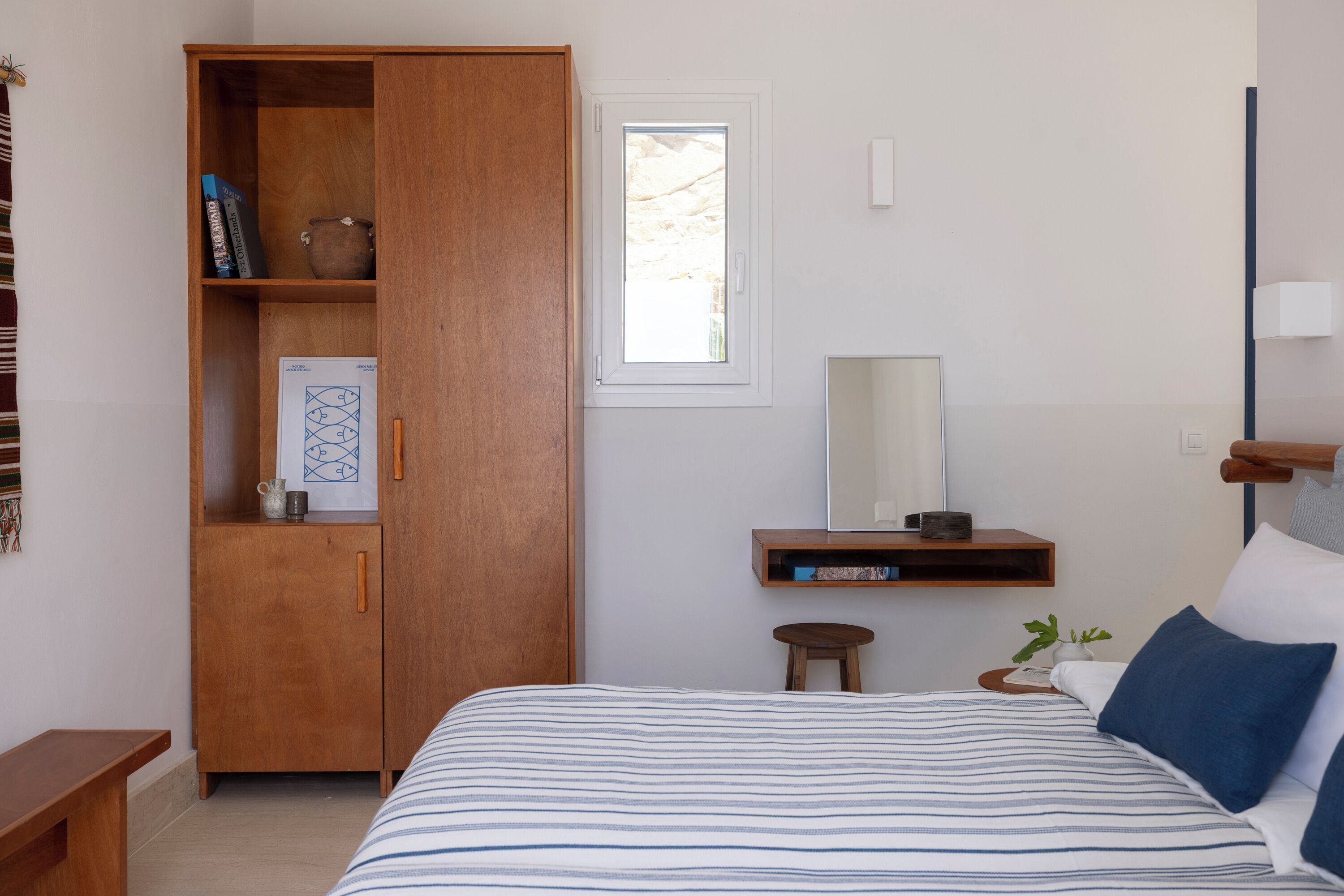 A bedroom with a bed, a wooden wardrobe, a small stool, a nightstand, a mirror, and a window.