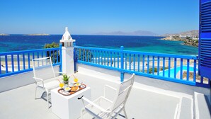 Un balcón con dos sillas blancas, una mesa pequeña con alimentos para el desayuno y vistas al mar y a la costa distante.