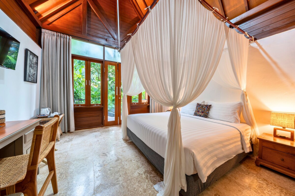 A bedroom with a canopy bed, a desk with a chair, and a view of greenery through the door.