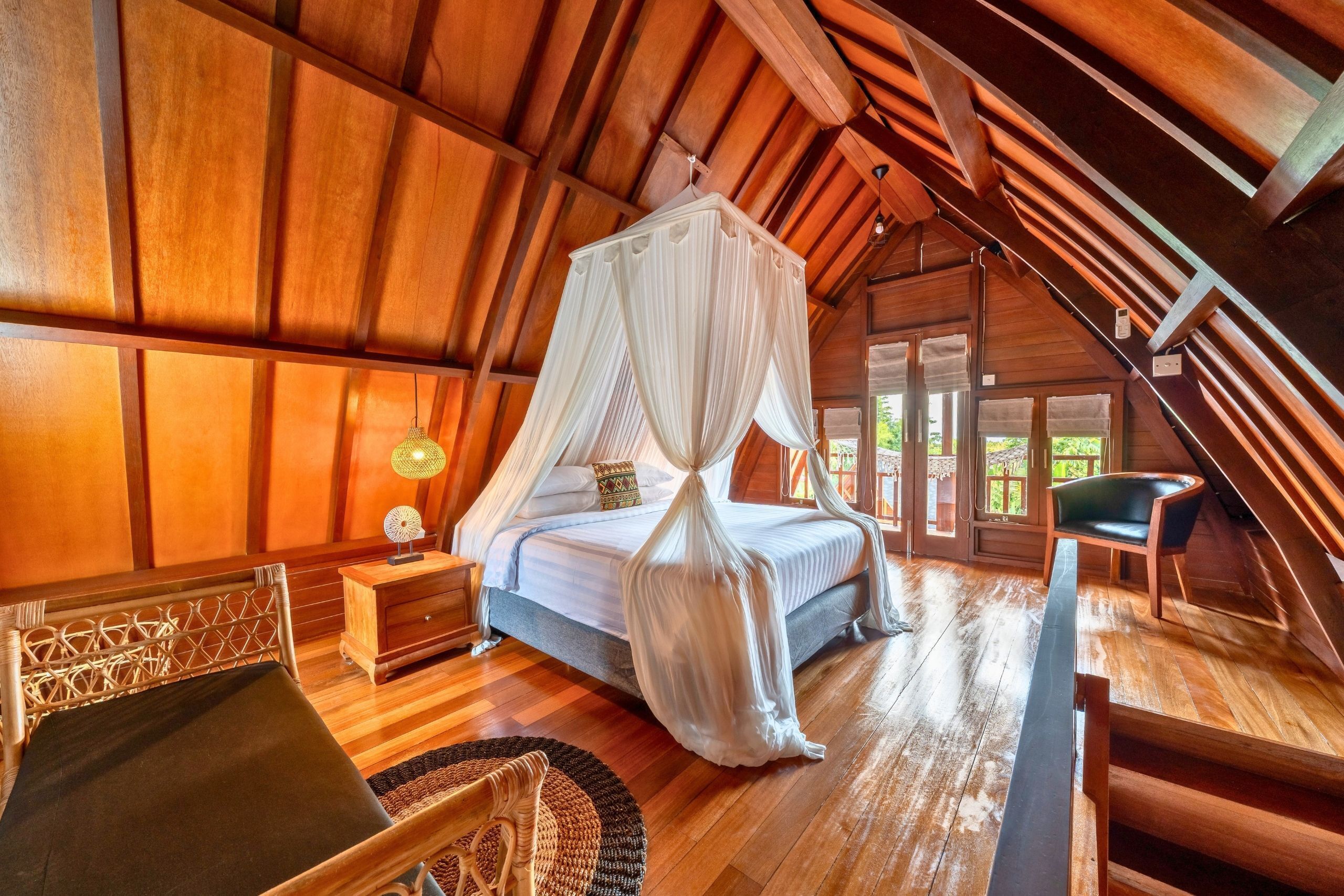 A bedroom with a canopy bed, wooden flooring, a nightstand, a chair, and a view of the outdoors.