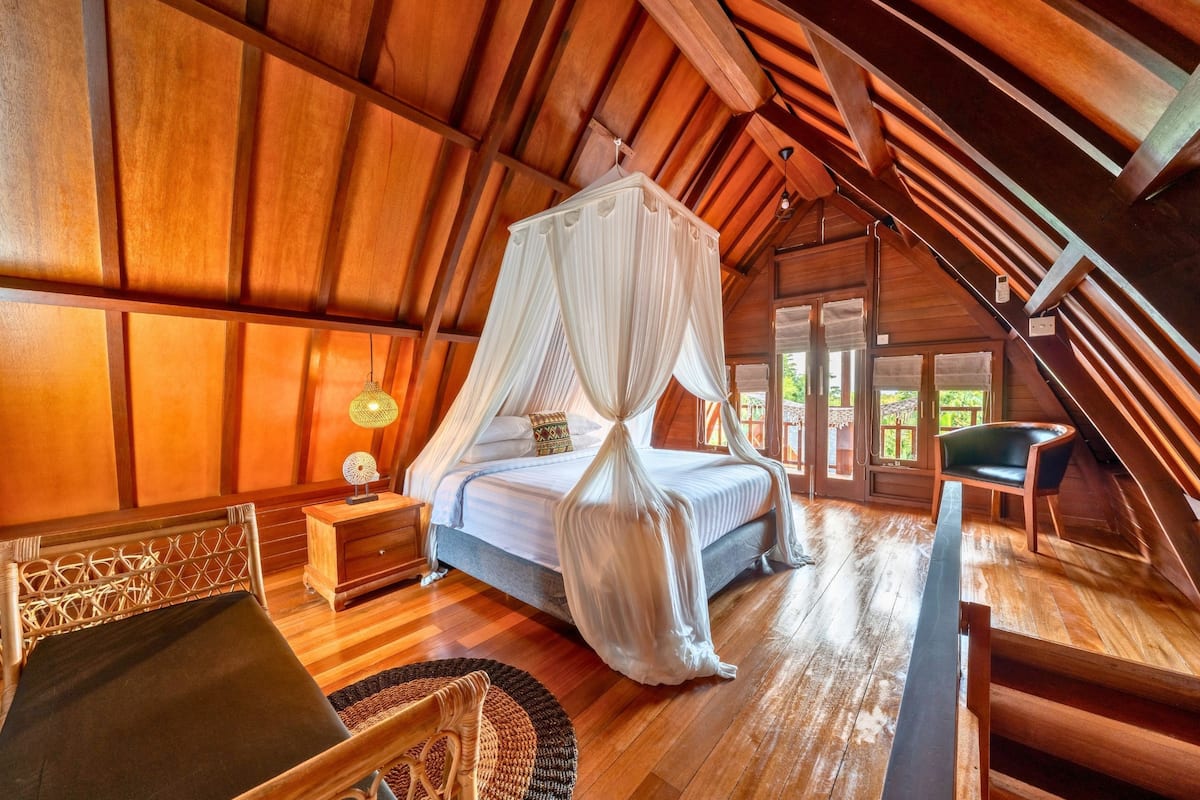 A bedroom with a canopy bed, wooden flooring, a nightstand, a chair, and a view of the outdoors.