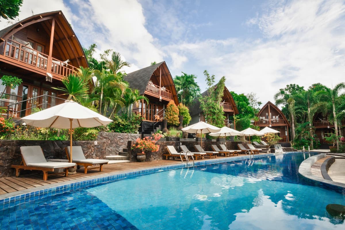 A pool area with wooden deck chairs and umbrellas, surrounded by lush greenery and wooden buildings.