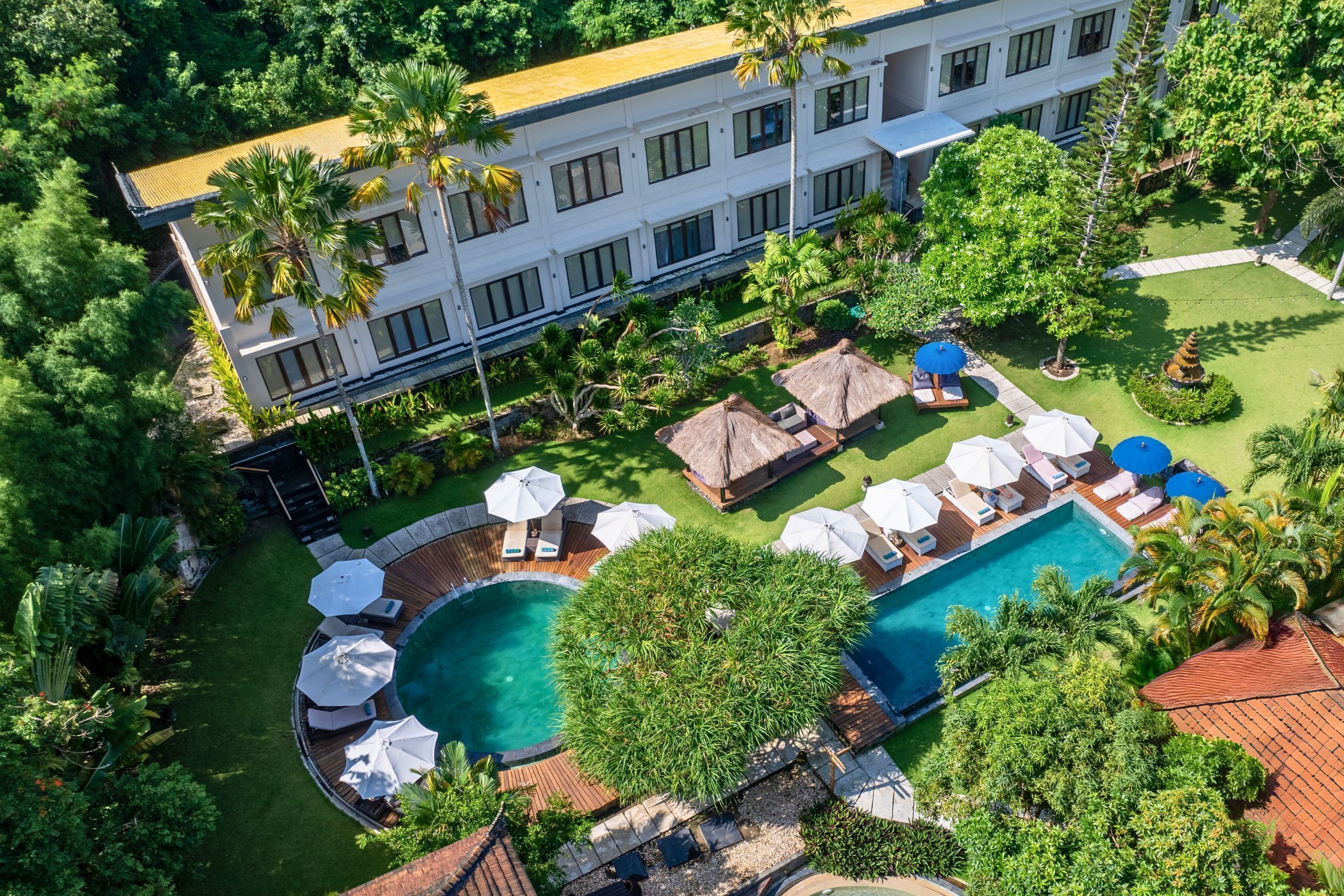 An aerial view of a resort with a swimming pool, sun loungers, umbrellas, and lush greenery.
