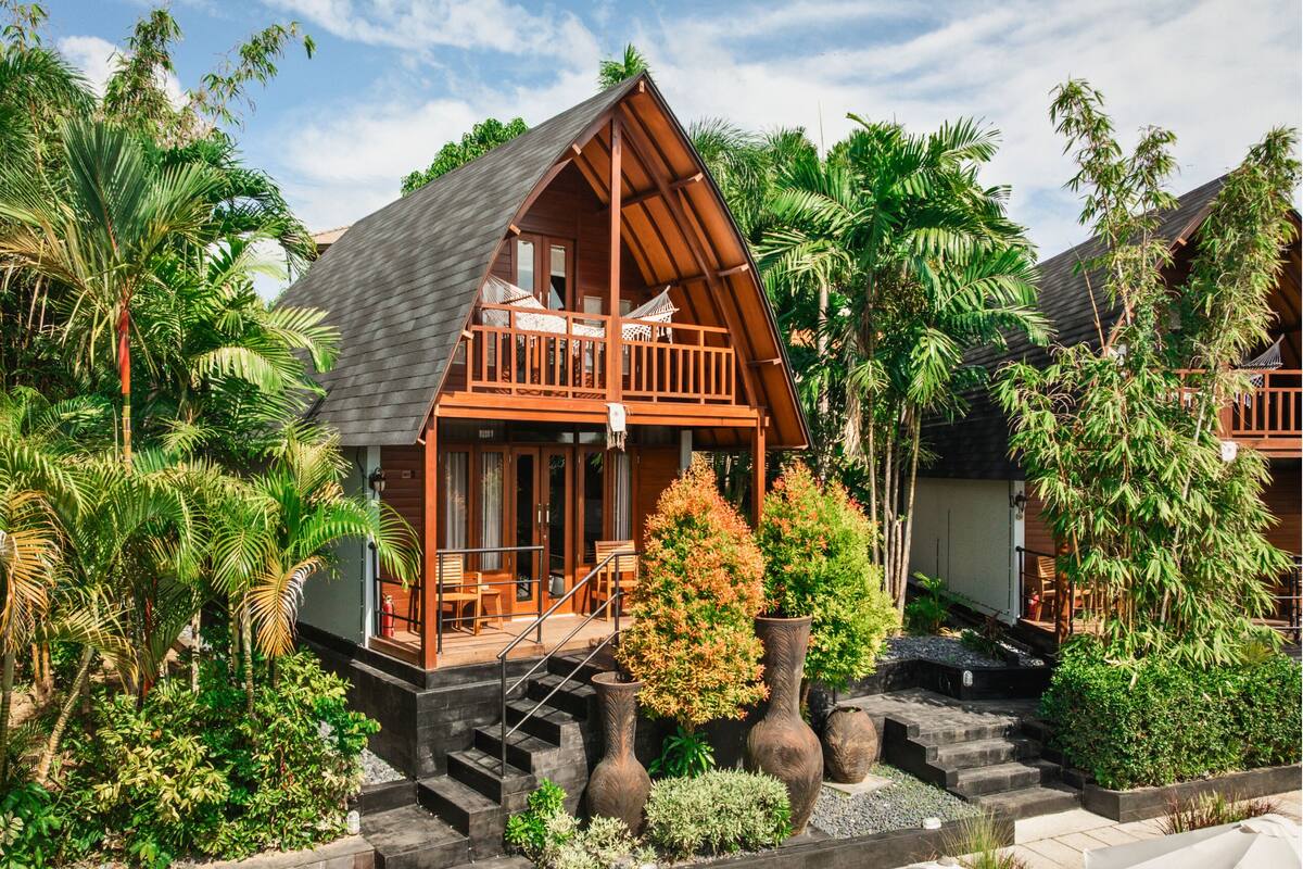 A wooden cabin with a balcony, surrounded by lush greenery and palm trees.