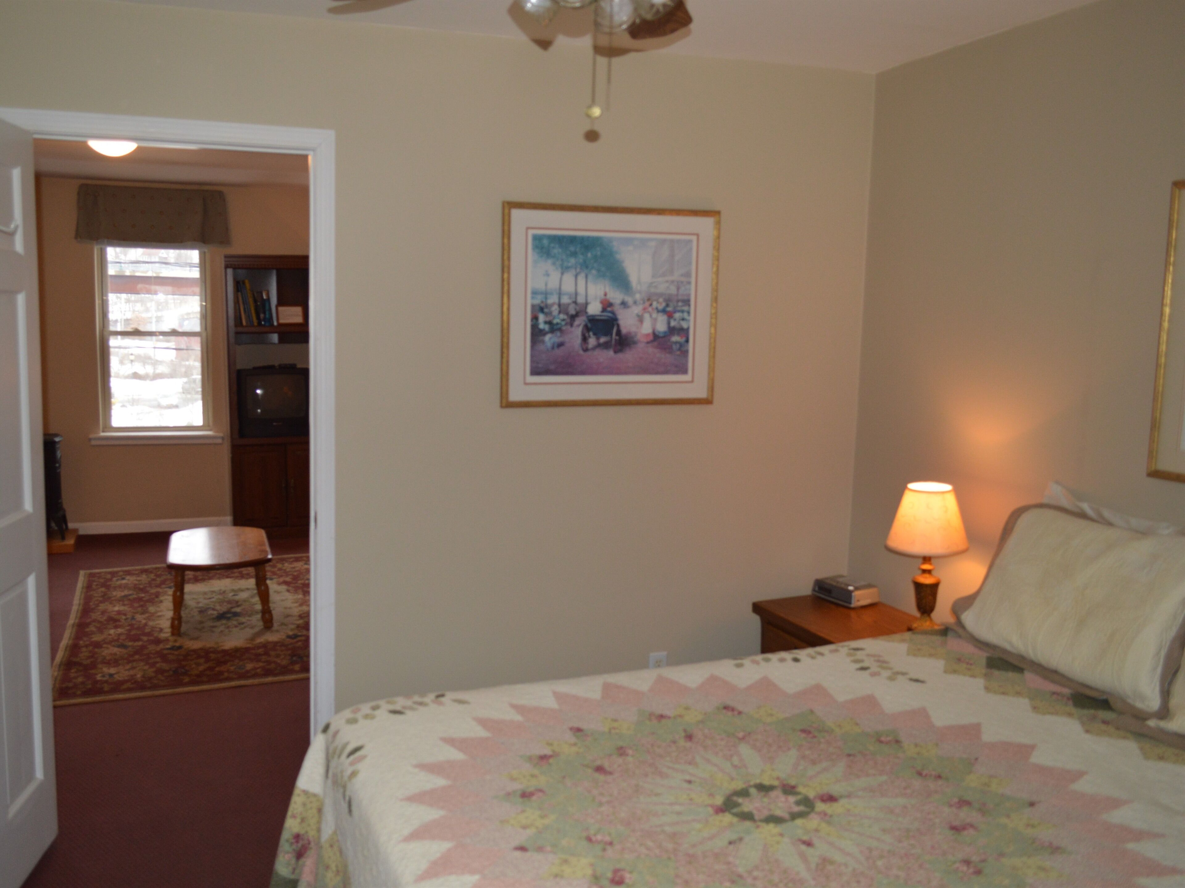 A bedroom with a bed, a nightstand with a lamp, a framed picture on the wall, a chair, and a desk with a computer.