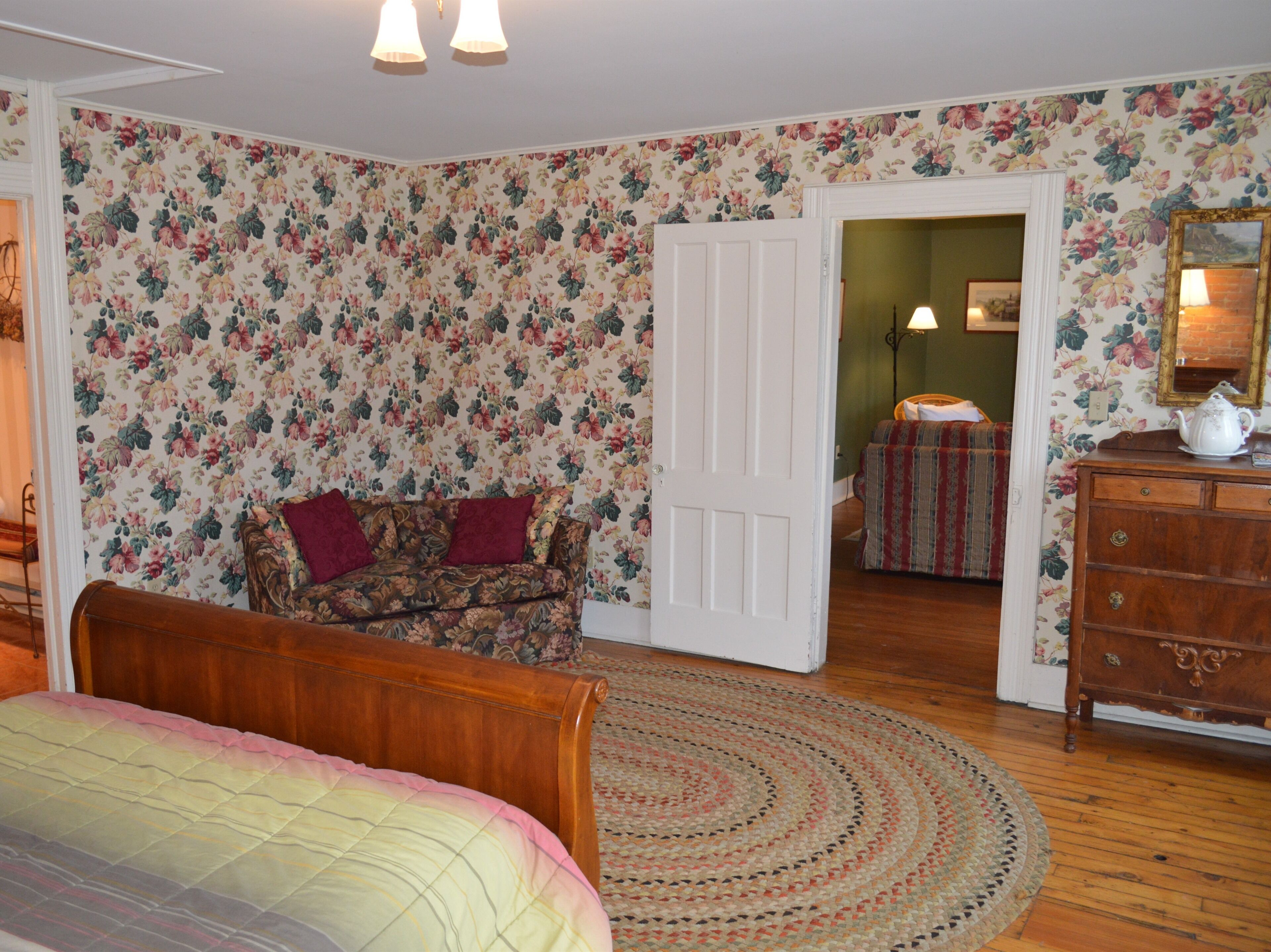 A bedroom with floral wallpaper, a wooden bed, a patterned sofa, a wooden dresser, and a round woven rug.