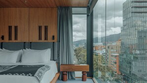 A modern hotel room with a large bed, a wooden bench, and a city view through large windows.