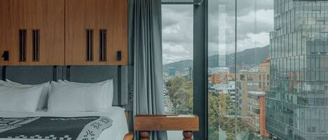 A modern hotel room with a large bed, a wooden bench, and a city view through large windows.