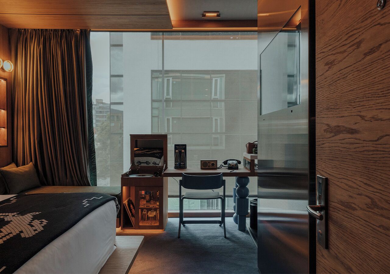 A modern hotel room with a bed, a desk with a chair, a small cabinet, and a large window offering a view of buildings outside.