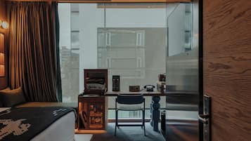 A modern hotel room with a bed, a desk with a chair, a small cabinet, and a large window offering a view of buildings outside.