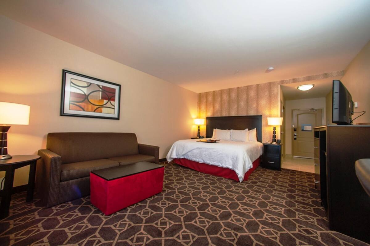 A hotel room with a bed, a sofa, a red ottoman, a TV, and a painting on the wall.