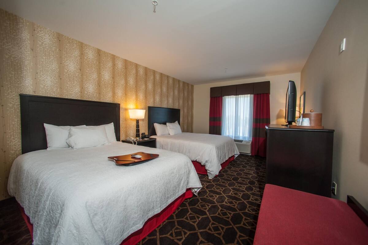 A hotel room with two beds, a desk, a lamp, a TV, and a red chair.