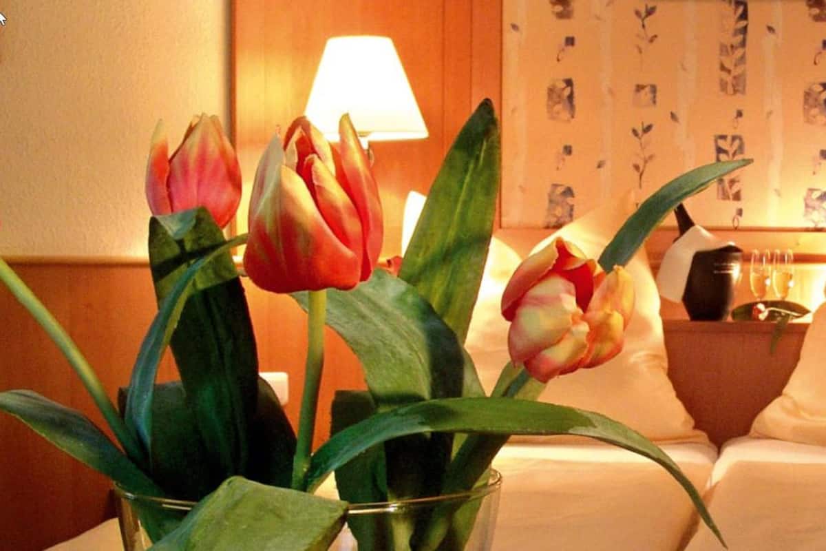 Quarto de hotel com uma cama, duas luminárias laterais, uma mesinha de cabeceira com luminária, um vaso com flores e uma parede com padrão floral.