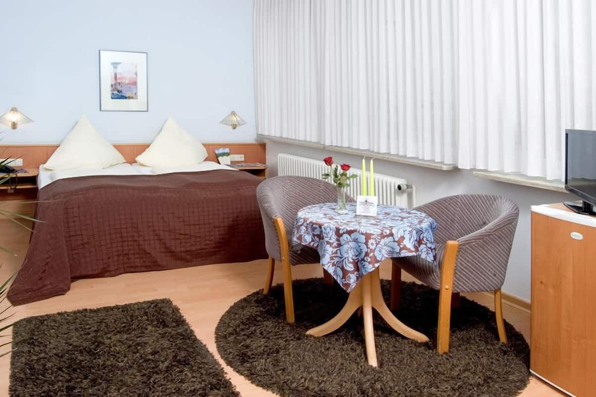 Quarto com cama, mesas de cabeceira, uma pequena mesa redonda com cadeiras, radiador e janela com cortinas.