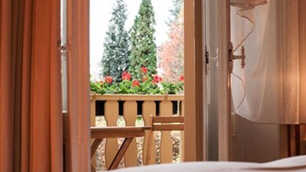 Ventana de dormitorio abierta con vistas a una terraza de madera y flores rojas.