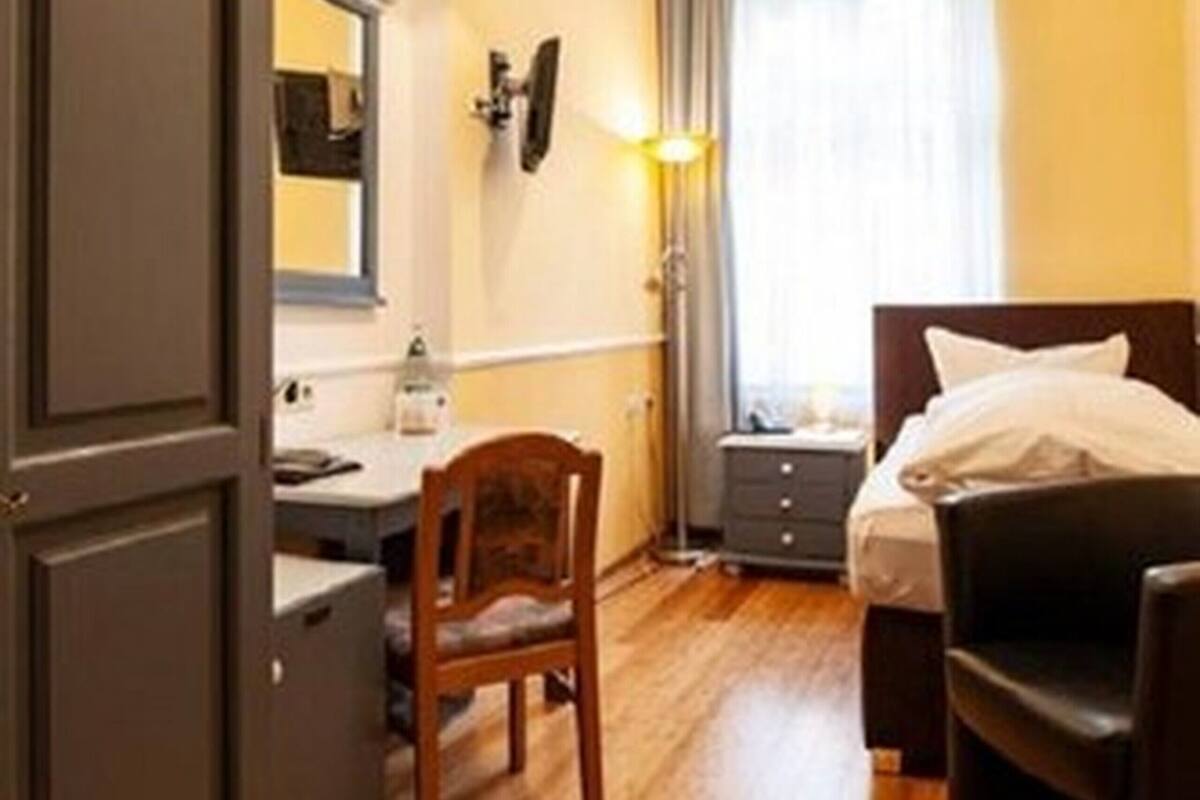 A hotel room with a wooden floor, a bed with white bedding, a desk with a chair, a wardrobe, and a window with curtains.