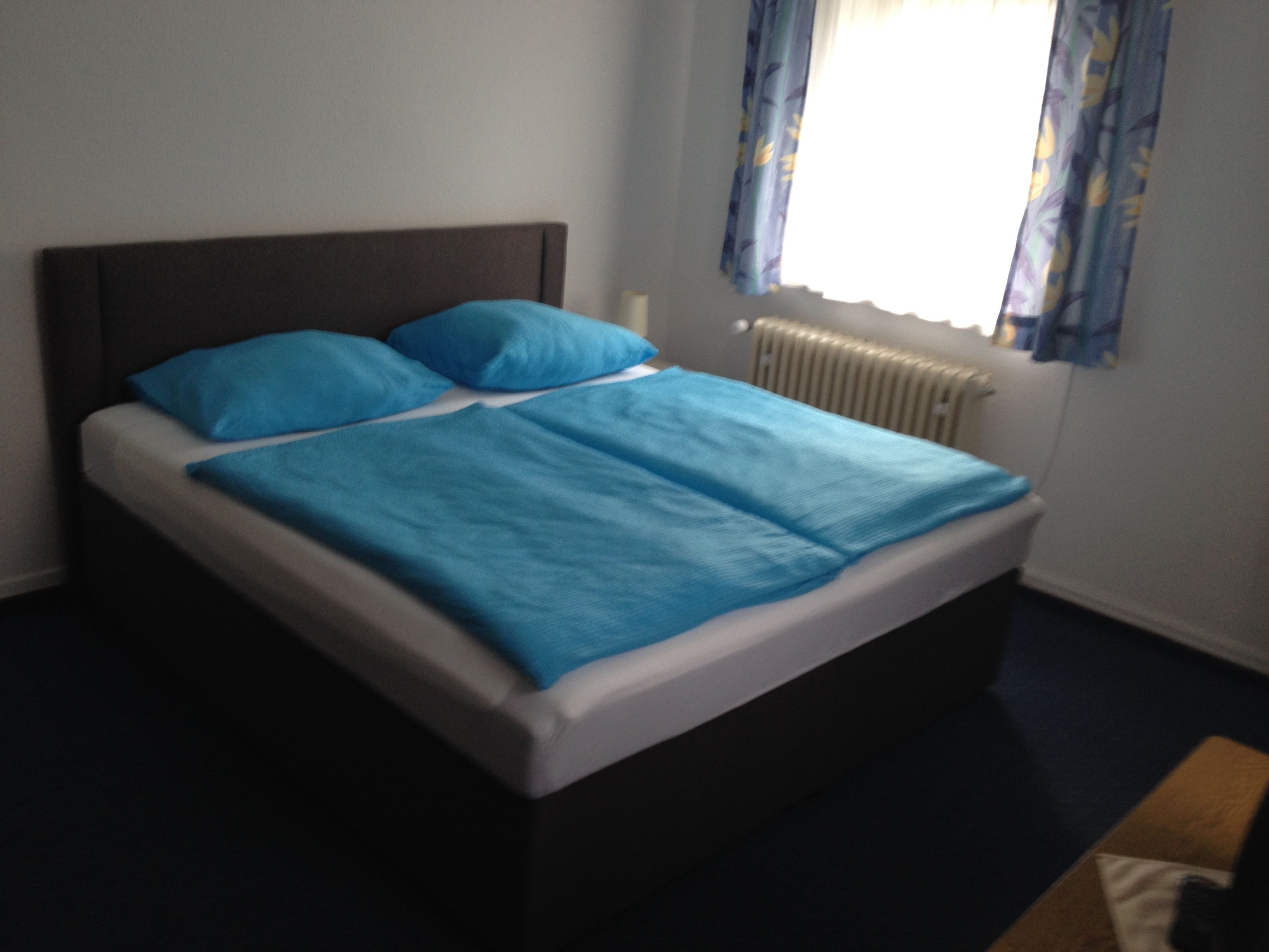 A bed with a blue bedspread and pillows, a white headboard, and a window with blue curtains.