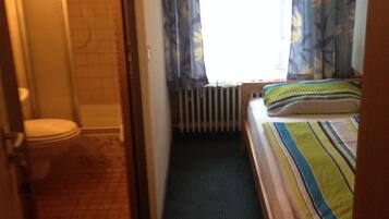 Una habitación de hotel pequeña y estrecha, con ducha, inodoro y una cama con manta a rayas.