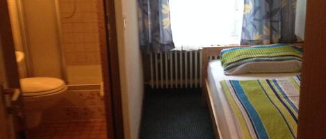Una habitación de hotel pequeña y estrecha, con ducha, inodoro y una cama con manta a rayas.
