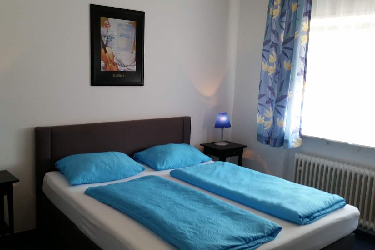 A neatly made bed with blue bedding, two bedside tables with lamps, a framed picture on the wall, and a window with curtains.