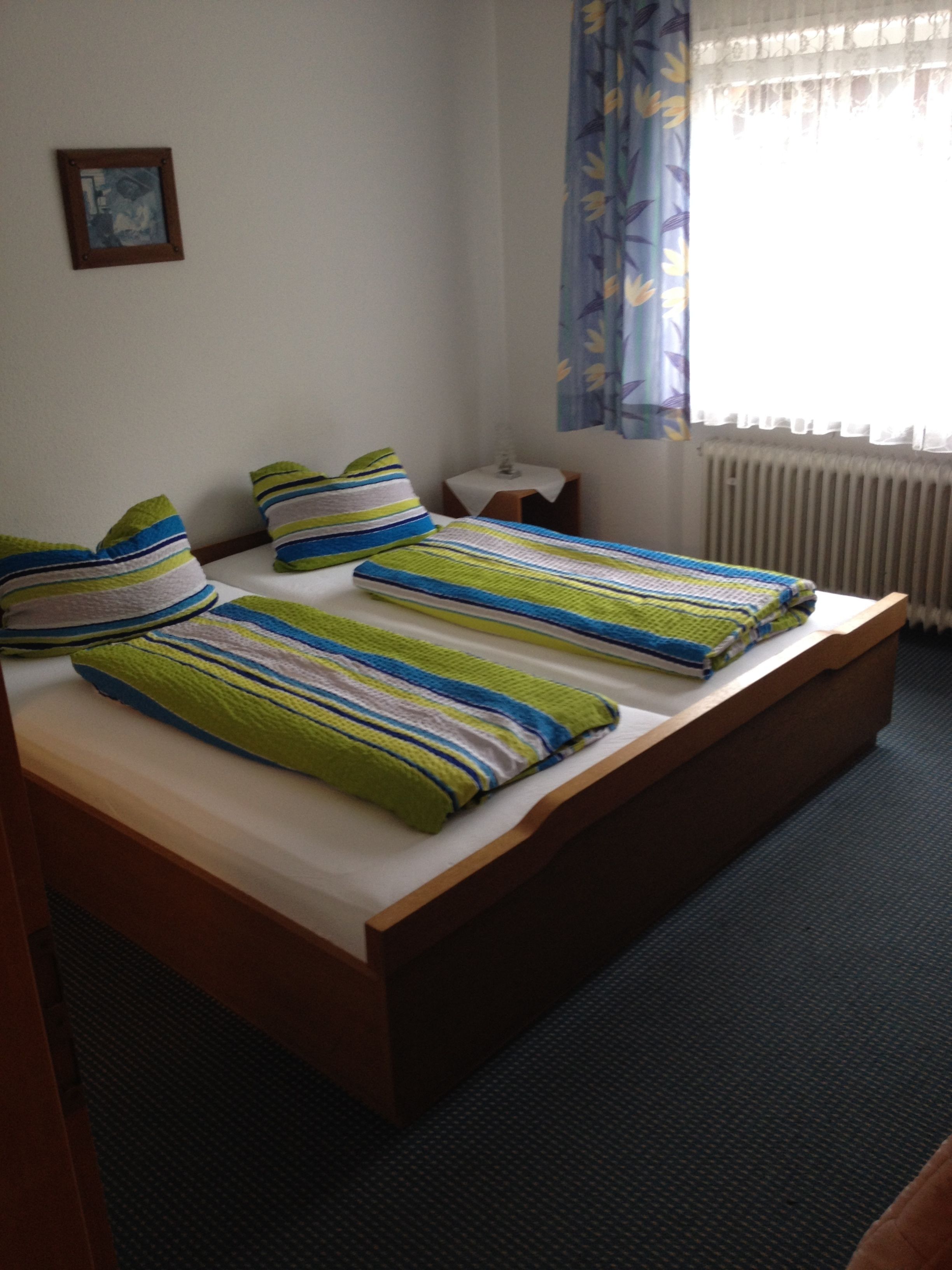 A double bed with striped bedding, a bedside table, a small nightstand, a radiator, and a window with curtains.