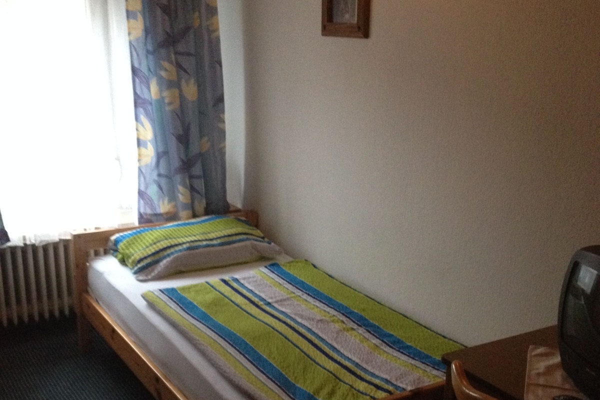 A single bed with a striped bedspread, a wooden nightstand, a television, and a radiator.