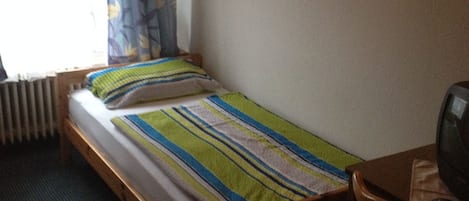 Una cama individual con colcha a rayas, una mesita de noche de madera, un televisor y un radiador.