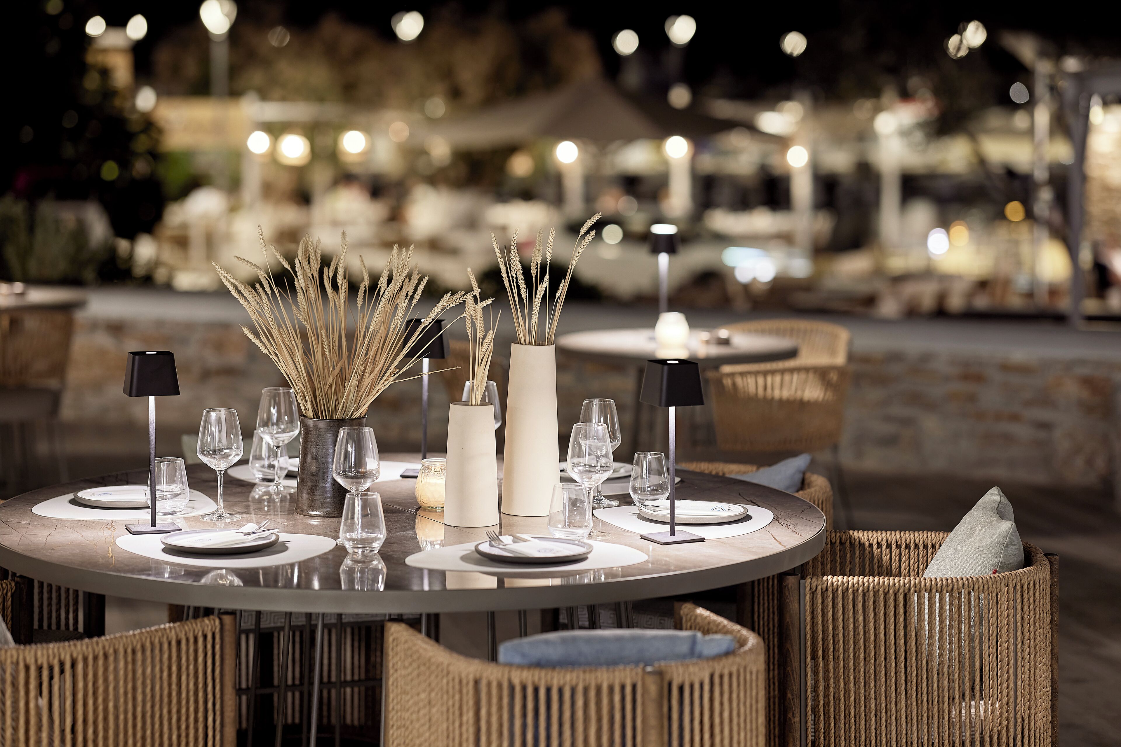 A round dining table set for four with candles, glasses, and a vase of dried plants.