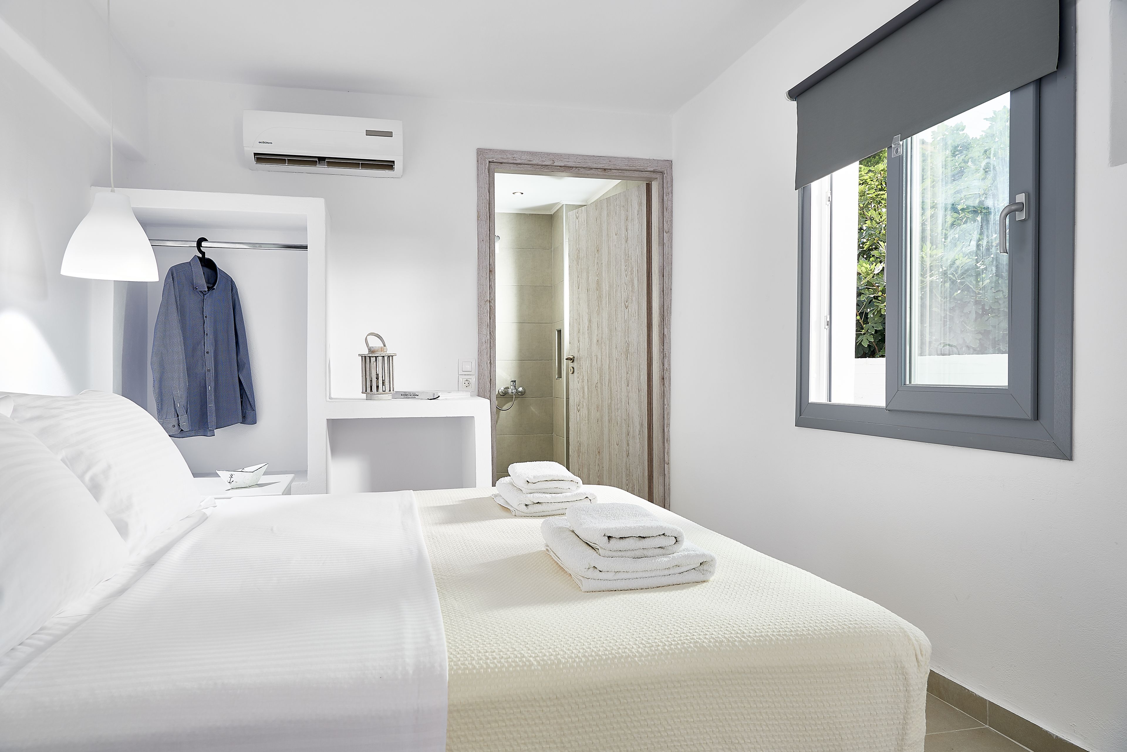 Una habitación de hotel moderna con una cama grande, una mesita de noche con una lámpara, un baño visible a través de una puerta abierta y una ventana con vistas a la vegetación.