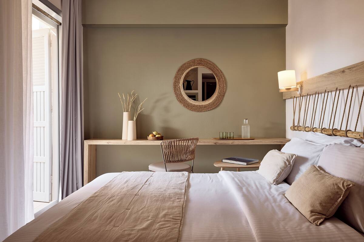 A bedroom with a bed, a desk with a chair, a mirror, and a bedside table with a lamp.