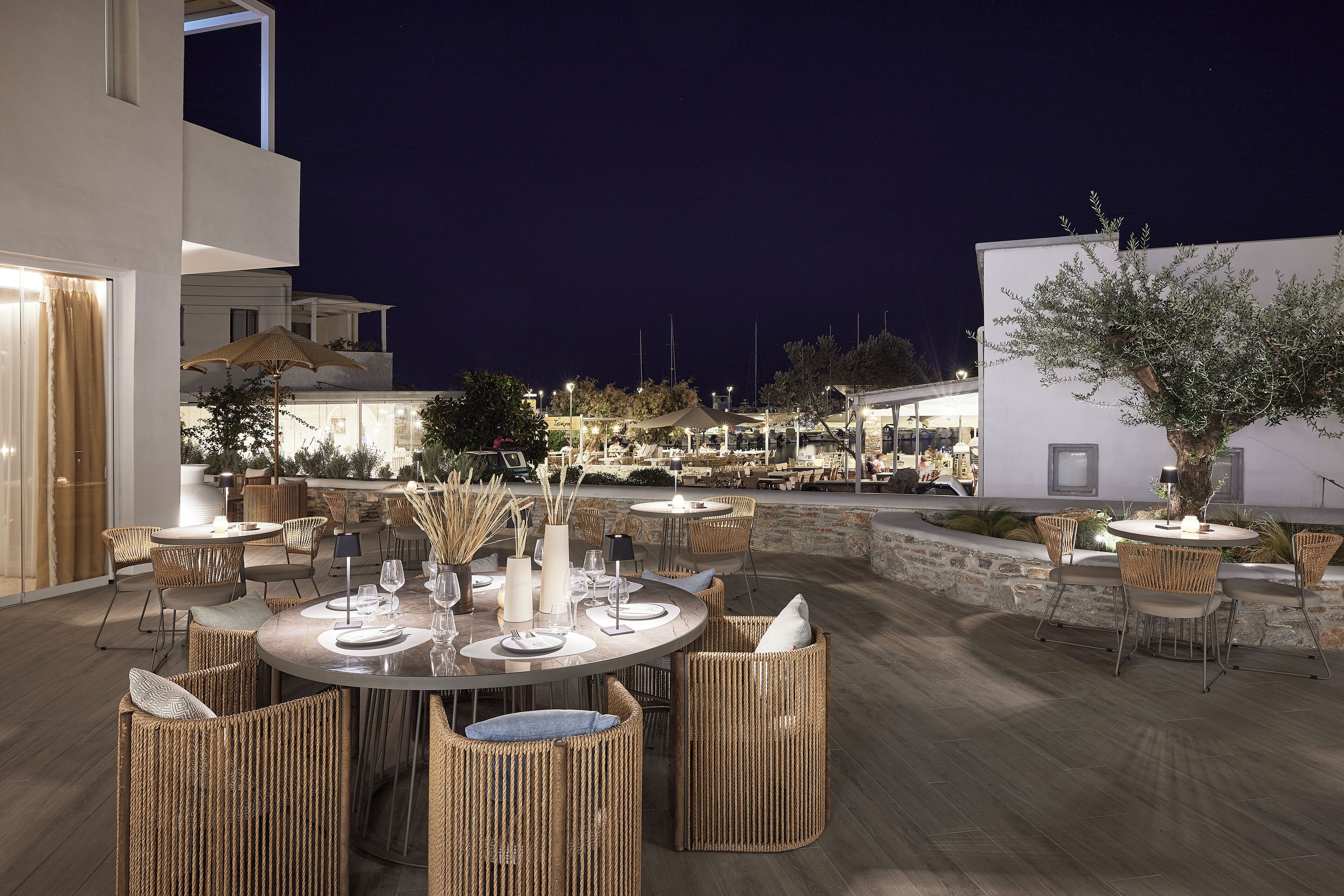 An outdoor dining area with round tables and wicker chairs, surrounded by a stone wall and a white building.