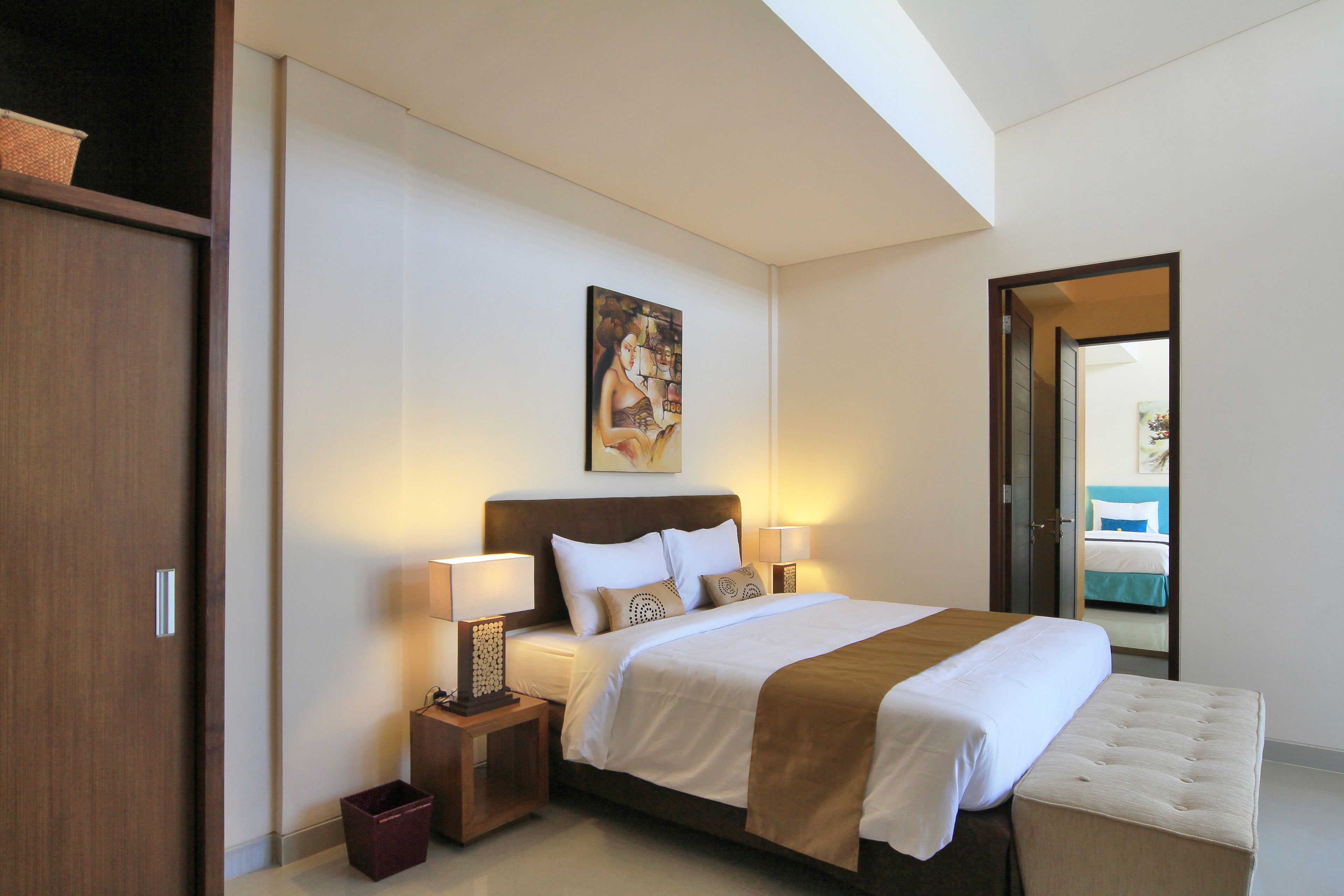 A hotel room with a large bed, two bedside tables with lamps, a wooden headboard, a painting above the bed, and a view of a pool through an open door.