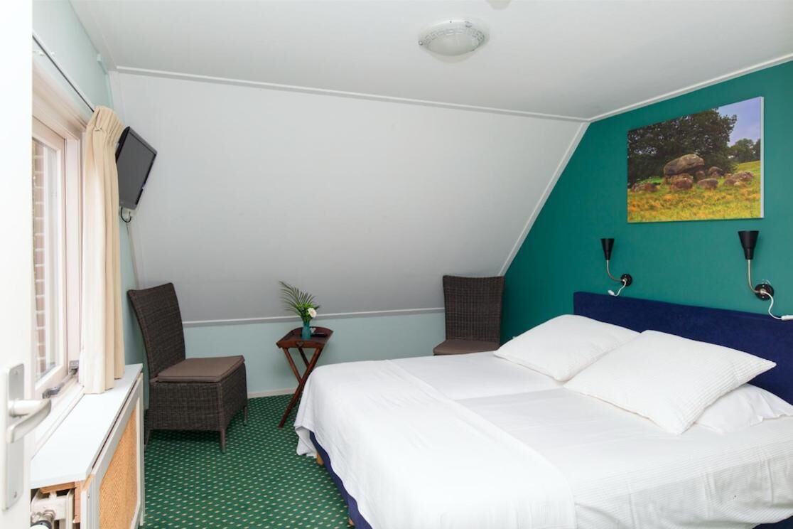 Een slaapkamer met een bed, twee stoelen, een tafeltje met een plant, een schilderij aan de muur en een raam met gordijnen.