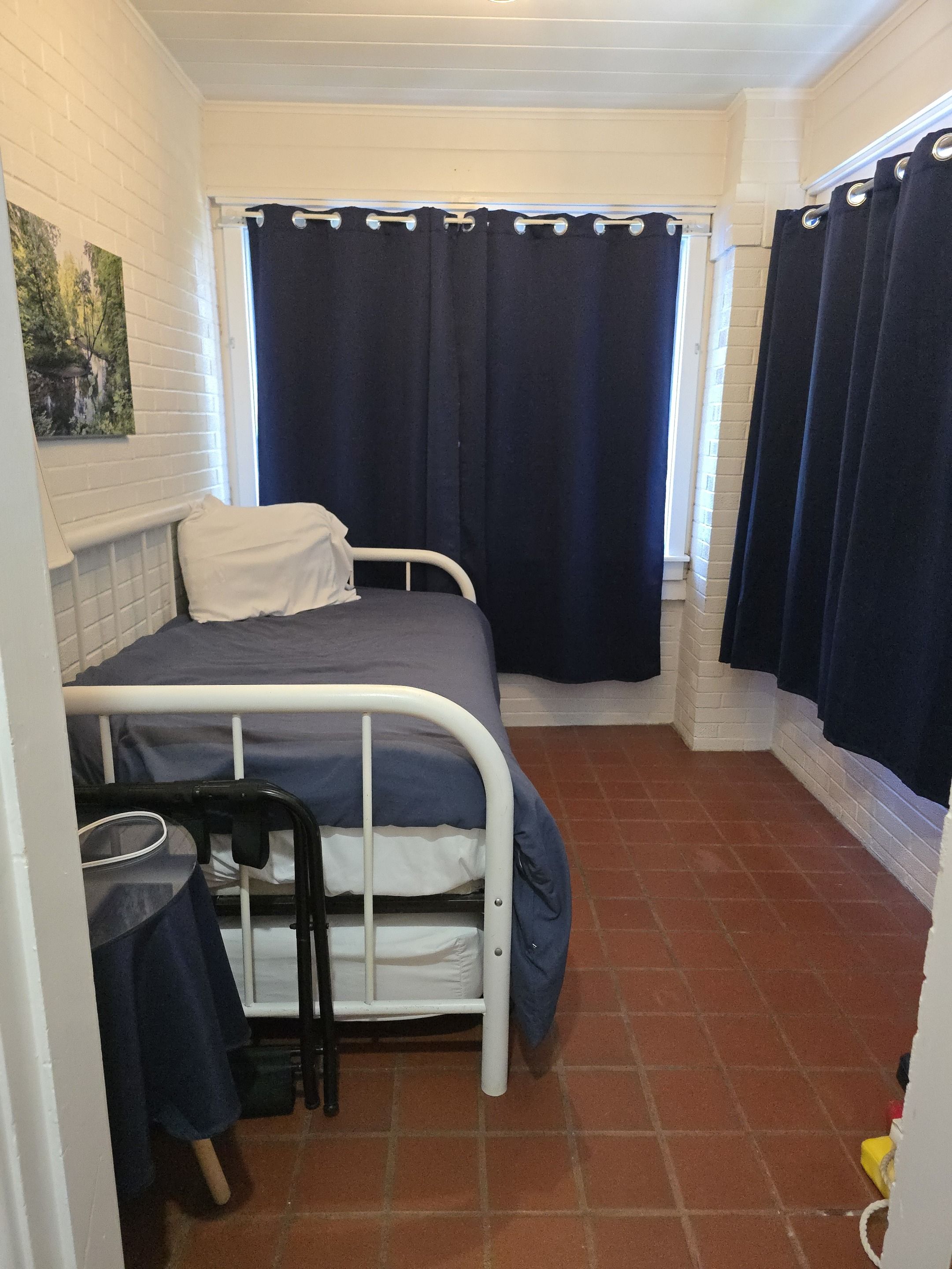 A small, single-bed room with a white metal bed frame, a blue mattress, and a white pillow. There is a small table with a chair, a window with dark curtains, and a tiled floor.