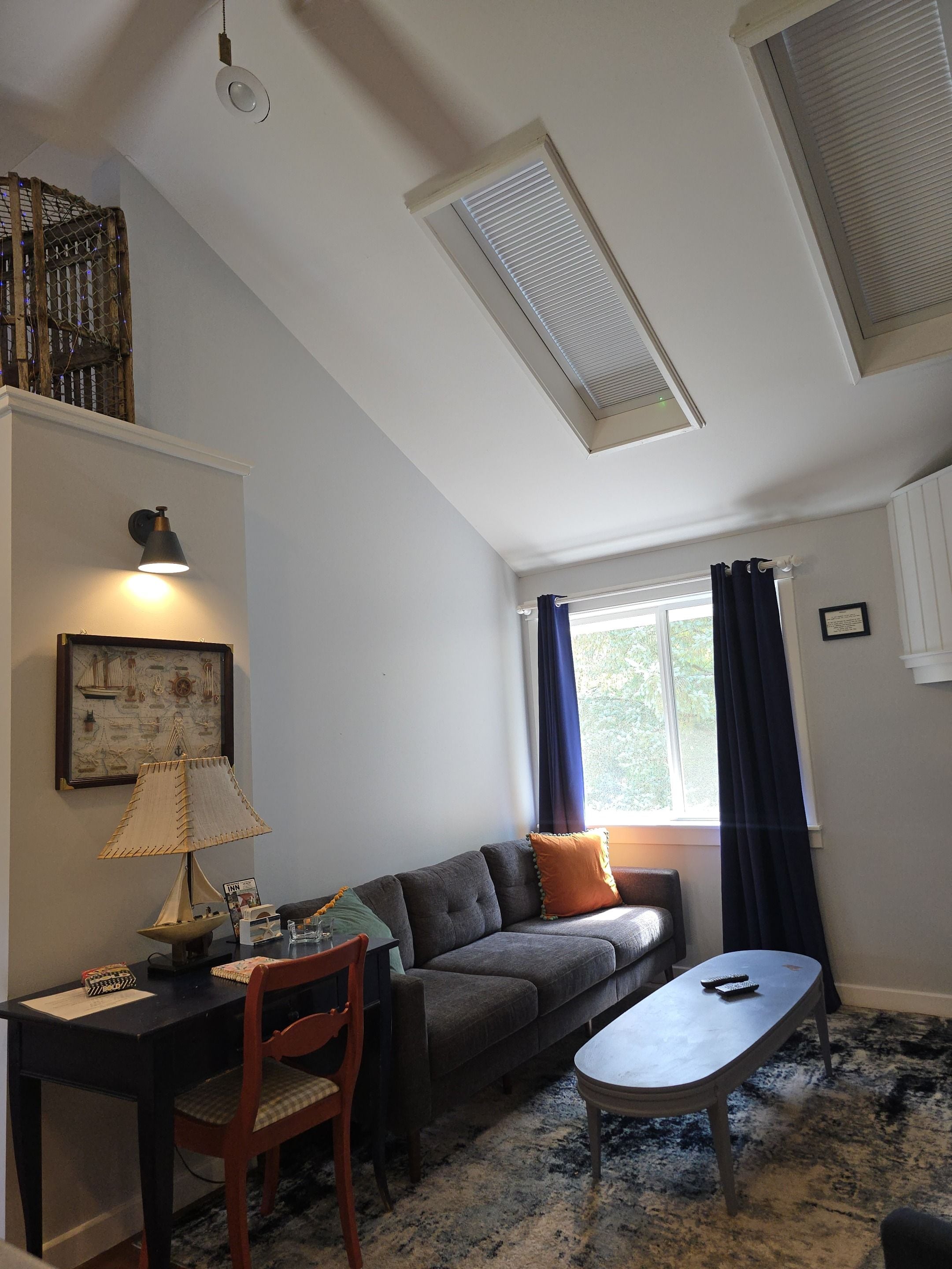 A living room with a sofa, a table, a chair, a lamp, a window with curtains, and a ceiling vent.
