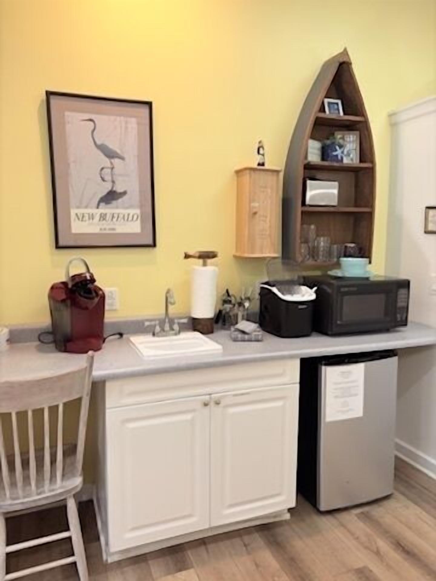 A compact kitchen with a white countertop, a sink, a microwave, and a small cabinet. There is a framed picture on the wall and a chair to the left.